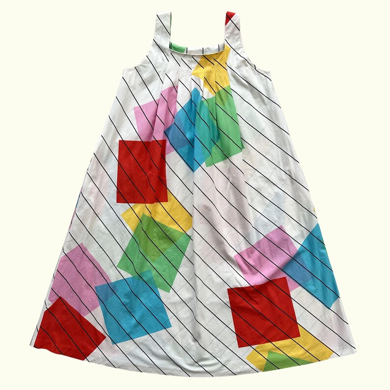 May include: White sleeveless dress with a colourful geometric pattern. The dress features large, vibrant squares in red, pink, blue, green, and yellow, set against a backdrop of black diagonal stripes. The straps have red and green accents.