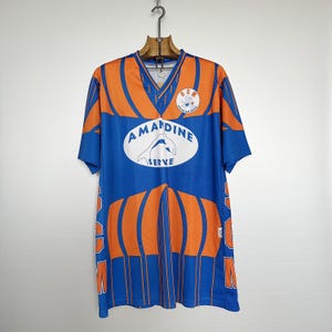 以下が含まれることがあります： 青とオレンジ色のヴィンテージサッカーユニフォーム、Vネック。ユニフォームには「AMANDINE SERVE」の文字と「SCM」の文字が入ったロゴが入っています。木製のハンガーに吊るされています。