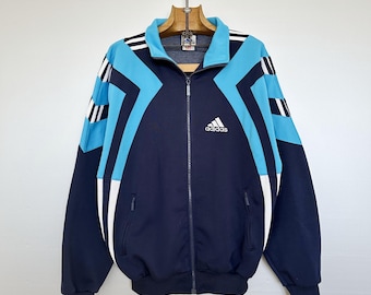May include: Navy blue and light blue Adidas track jacket with white accents. The jacket features a full zip, two front pockets, and the Adidas logo on the chest. The design includes geometric patterns and stripes on the sleeves.