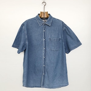 Puede incluir: Camisa vaquera azul claro de manga corta con botones y un bolsillo en el pecho. La camisa est&aacute; colgada de una percha de madera sobre un fondo blanco. La camisa tiene un cuello cl&aacute;sico.