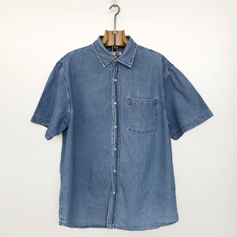 Puede incluir: Camisa vaquera azul claro de manga corta con botones y un bolsillo en el pecho. La camisa est&aacute; colgada de una percha de madera sobre un fondo blanco. La camisa tiene un cuello cl&aacute;sico.