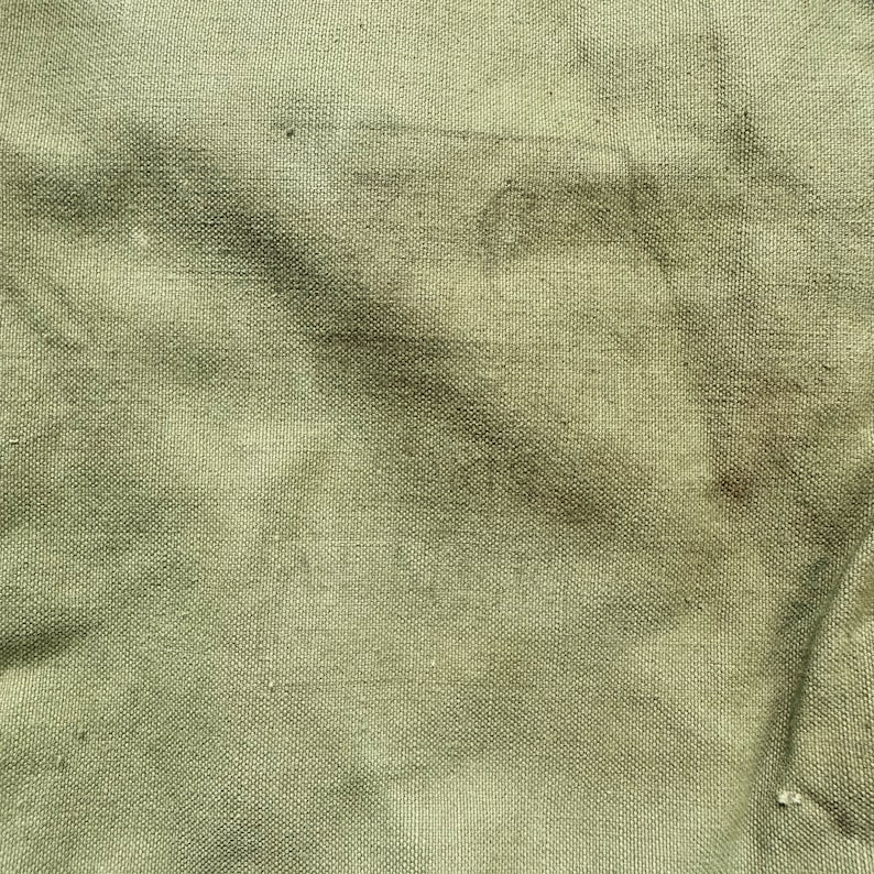 May include: A close-up of a green canvas fabric with a textured weave. The fabric has a few dark brown stains.