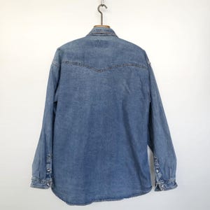 Pode incluir: Camisa jeans azul clara com mangas compridas, punhos abotoados e gola cl&aacute;ssica. A parte de tr&aacute;s da camisa apresenta um detalhe de carneira. A camisa est&aacute; pendurada num gancho de metal sobre um fundo branco.