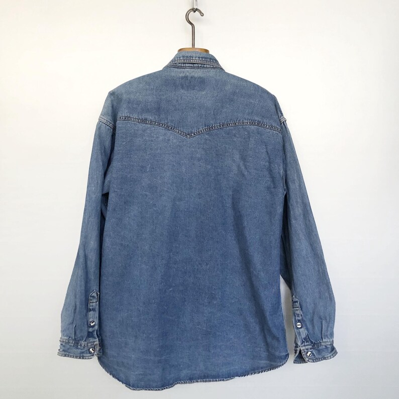 Pode incluir: Camisa jeans azul clara com mangas compridas, punhos abotoados e gola cl&aacute;ssica. A parte de tr&aacute;s da camisa apresenta um detalhe de carneira. A camisa est&aacute; pendurada num gancho de metal sobre um fundo branco.