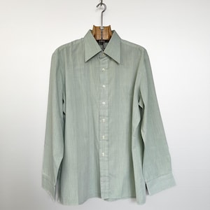 Könnte beinhalten: Hellgrünes Hemd mit langen Ärmeln, das an einem Holzkleiderbügel hängt. Das Hemd hat ein vertikales Streifenmuster und einen klassischen Kragen. Es ist zugeknöpft und scheint aus leichtem Stoff zu sein.
