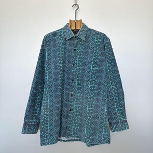 May include: A long-sleeved button-down shirt with a teal and navy geometric pattern. The shirt is hanging from a wooden hanger. The pattern is made up of repeating shapes and designs. The shirt has a collar and buttoned cuffs.