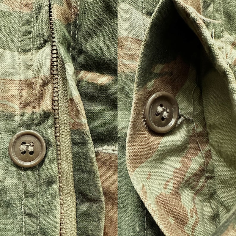 May include: Close-up of a green, brown, and tan camouflage fabric with a zipper and two brown buttons. The fabric is textured and has a worn look.