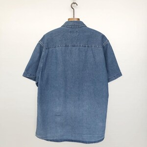 Puede incluir: Camisa vaquera de manga corta en un lavado azul medio, colgada de una percha de madera. La camisa presenta un cuello cl&aacute;sico, una parte delantera abotonada y un detalle de canes&uacute; en la espalda. La tela parece ser un denim de algod&oacute;n resistente.