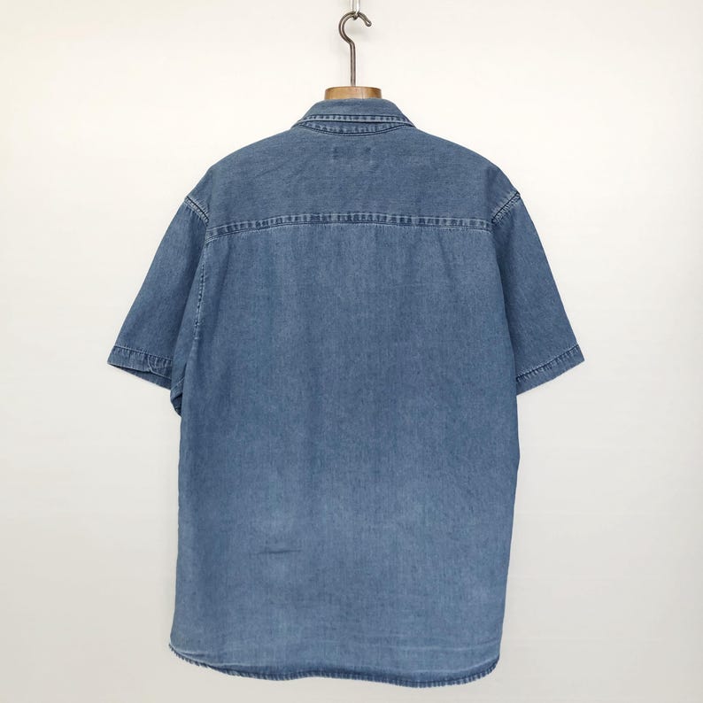 Puede incluir: Camisa vaquera de manga corta en un lavado azul medio, colgada de una percha de madera. La camisa presenta un cuello cl&aacute;sico, una parte delantera abotonada y un detalle de canes&uacute; en la espalda. La tela parece ser un denim de algod&oacute;n resistente.