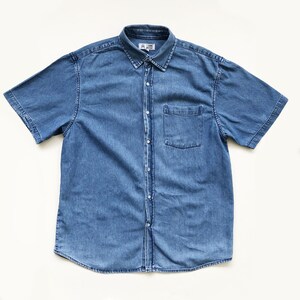 Puede incluir: Una camisa de mezclilla azul claro de manga corta con botones en la parte delantera y un bolsillo en el pecho. La camisa tiene un cuello cl&aacute;sico y est&aacute; hecha de una tela de mezclilla de peso medio. La camisa est&aacute; tendida sobre una superficie blanca.