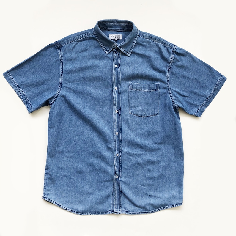 Puede incluir: Una camisa de mezclilla azul claro de manga corta con botones en la parte delantera y un bolsillo en el pecho. La camisa tiene un cuello cl&aacute;sico y est&aacute; hecha de una tela de mezclilla de peso medio. La camisa est&aacute; tendida sobre una superficie blanca.