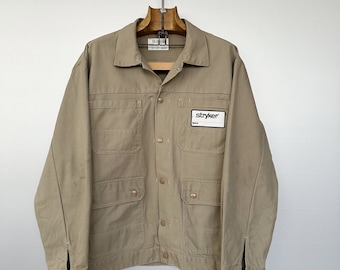 Può includere: Una giacca da lavoro beige con chiusura a bottoni e diverse tasche. La giacca ha una toppa sul petto sinistro che recita "stryker".