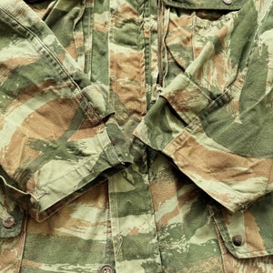May include: Close-up of a camouflage jacket with a zipper closure. The jacket is made of a brown, green, and tan fabric with a pattern of stripes and swirls. The fabric is textured and has a worn look.