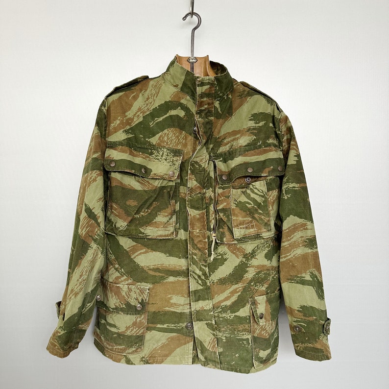 May include: A green and brown camouflage jacket with a zipper and button closure. The jacket has multiple pockets and a drawstring hood.