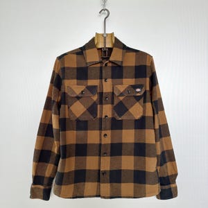 Puede incluir: Una camisa de franela a cuadros marrón y negro con dos bolsillos en el pecho y un pequeño logotipo en el bolsillo derecho. La camisa es abotonada y cuelga de una percha de madera.