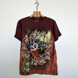 May include: Maroon short-sleeve t-shirt with a tattoo-style graphic. The design features a blue swallow, roses, a heart, and the text "Emperor for Eternity" in gold script. The shirt is hanging on a wooden hanger.
