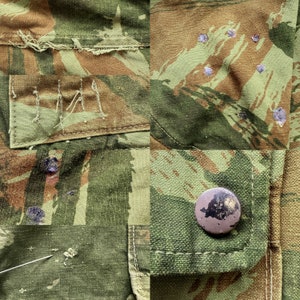 May include: Close-up of a camouflage fabric with a brown button and a patch with the letters "IMA" stitched on it. The fabric has a worn and faded appearance with several small holes.