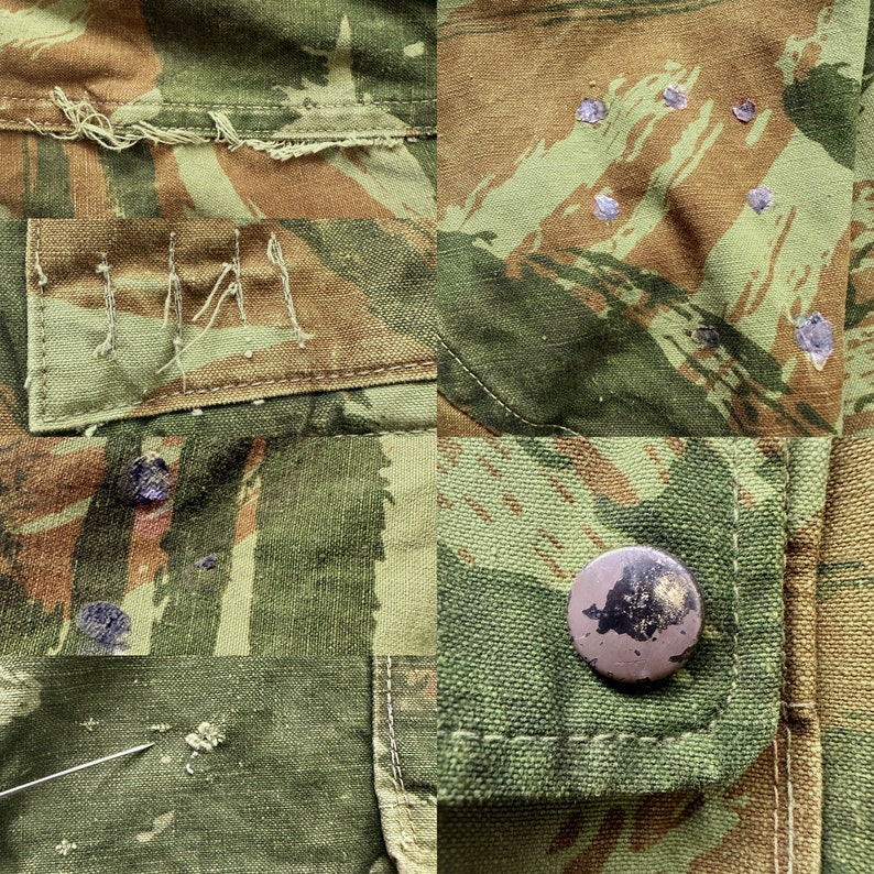 May include: Close-up of a camouflage fabric with a brown button and a patch with the letters "IMA" stitched on it. The fabric has a worn and faded appearance with several small holes.