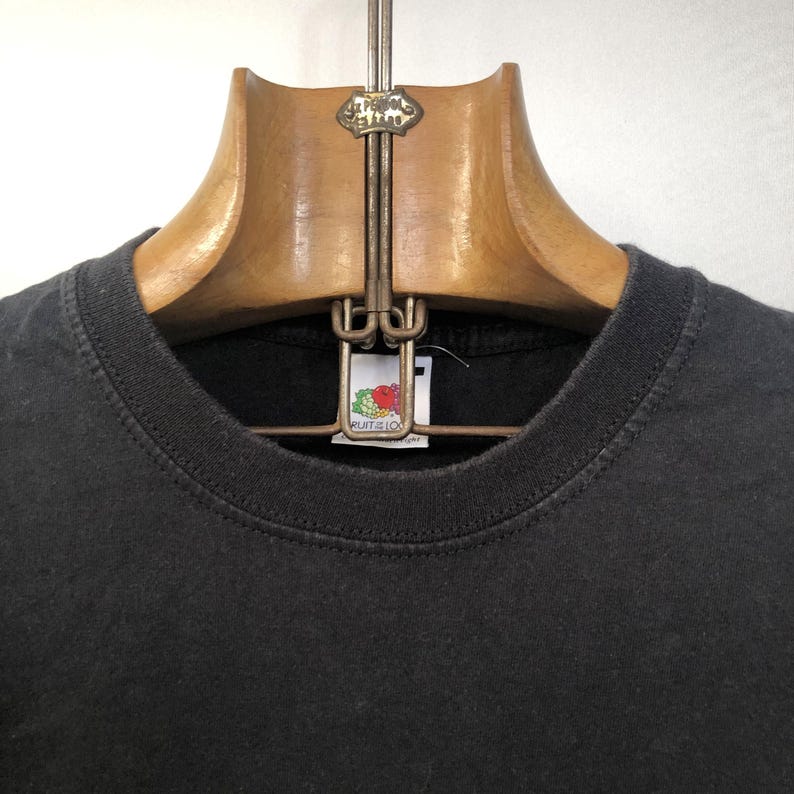 K&ouml;nnte beinhalten: Schwarzes T-Shirt mit einem Fruit of the Loom-Etikett. Das Shirt h&auml;ngt an einem Holzb&uuml;gel.
