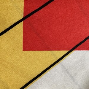 May include: Close-up of a textile with an abstract design. It features a red square, yellow and white triangles, and black diagonal lines. The fabric has a textured appearance, with a focus on the geometric shapes and colour blocking. The design is reminiscent of modern art.