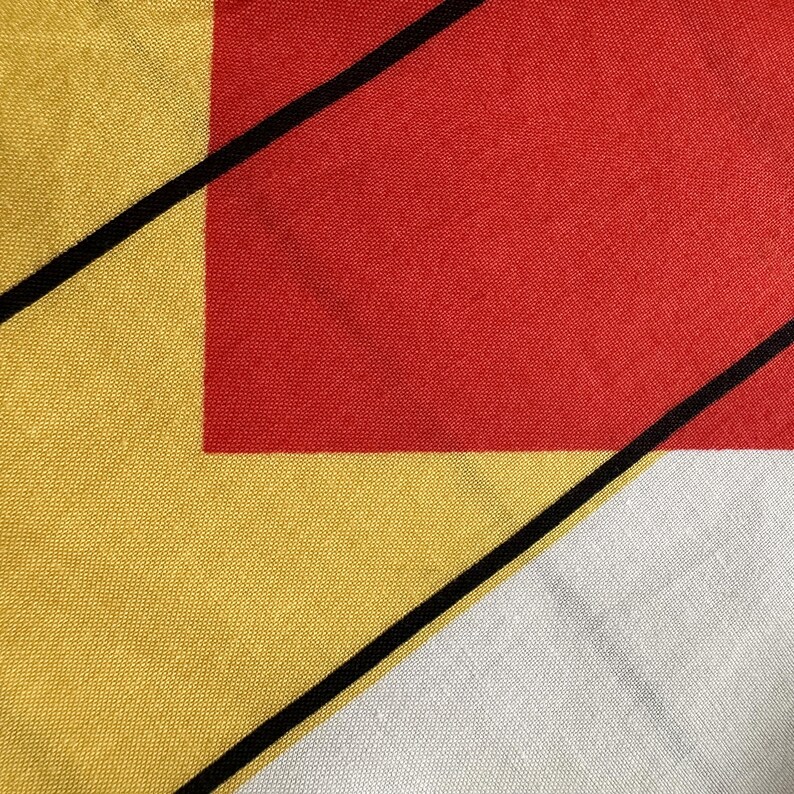 May include: Close-up of a textile with an abstract design. It features a red square, yellow and white triangles, and black diagonal lines. The fabric has a textured appearance, with a focus on the geometric shapes and colour blocking. The design is reminiscent of modern art.