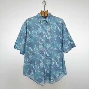May include: A short-sleeved button-down shirt with a teal and blue floral and geometric pattern. The shirt is hanging on a wooden hanger against a white background. The shirt has a classic collar and buttoned front.