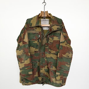 May include: Camouflage jacket in shades of green, brown, and tan. The jacket features multiple pockets, a zipper closure, and button details. It is hanging on a wooden hanger.