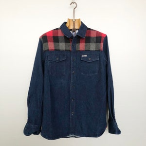 Peut inclure: Une chemise en jean bleu à manches longues avec un empiècement en écossais rouge et noir. La chemise a un devant boutonné et deux poches poitrine.