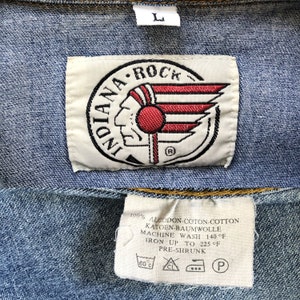 Pode incluir: Close-up de uma jaqueta jeans azul com uma etiqueta branca com o texto "INDIANA ROCK" e um logotipo de cabe&ccedil;a nativa americana com detalhes em vermelho e branco. Uma etiqueta menor abaixo fornece instru&ccedil;&otilde;es de cuidados.