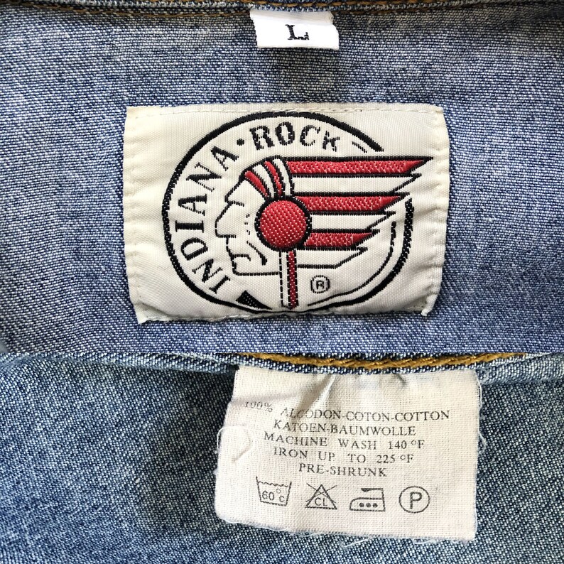 Pode incluir: Close-up de uma jaqueta jeans azul com uma etiqueta branca com o texto "INDIANA ROCK" e um logotipo de cabe&ccedil;a nativa americana com detalhes em vermelho e branco. Uma etiqueta menor abaixo fornece instru&ccedil;&otilde;es de cuidados.