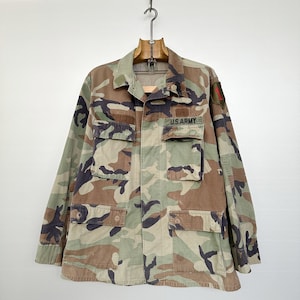 Może przedstawiać: Kamuflażowa kurtka wojskowa z naszywką U.S. Army na lewej piersi i czerwoną naszywką na prawym rękawie.