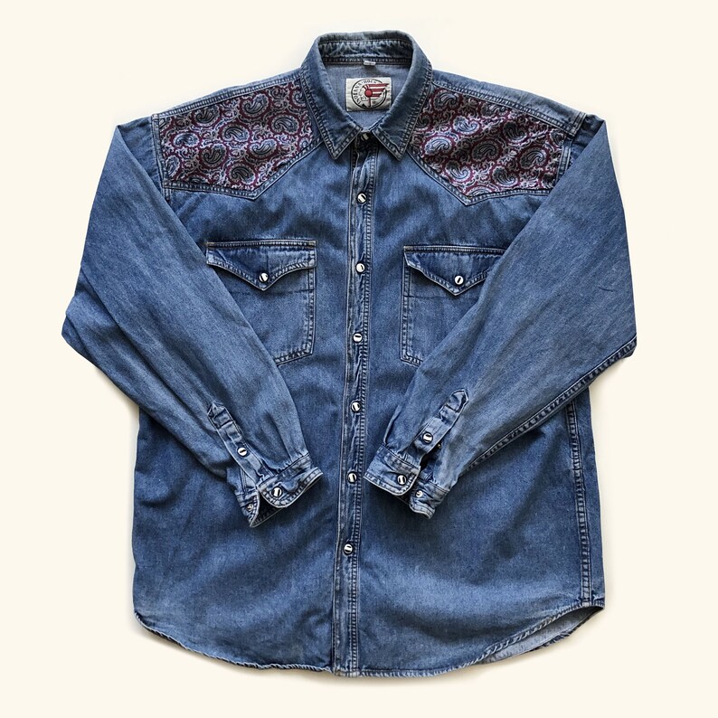 Pode incluir: Camisa jeans azul com mangas compridas e fecho de bot&otilde;es. A camisa apresenta um padr&atilde;o paisley em tons de bord&ocirc;, azul e branco nos ombros. Possui dois bolsos no peito e punhos abotoados.