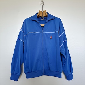 Puede incluir: Chaqueta deportiva azul brillante con cremallera y detalles blancos. Presenta cuello alzado, puños y dobladillo de canalé. Un pequeño emblema es visible. La chaqueta está colgada de una percha de madera.