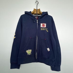 May include: Navy blue zip-up hoodie with a hood and front pockets. Features embroidered patches including a red dragon, a crown, and text. The sleeves have a yellow design. The hoodie is hanging on a wooden hanger.
