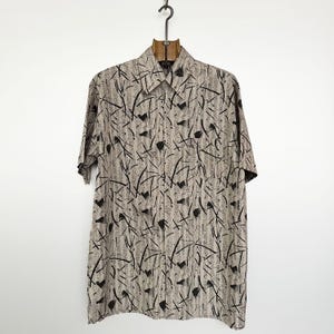 Puede incluir: Una camisa de manga corta con botones, con un fondo beige y un patrón abstracto negro. La camisa tiene cuello, un bolsillo en el pecho y está colgada de una percha de metal negro.