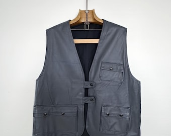 May include: A grey leather vest with two front pockets and two button closures. The sleeveless vest hangs on a wooden hanger against a white background. The vest has a black interior lining.