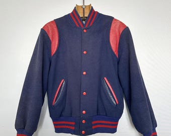 May include: A vintage navy blue varsity jacket with red leather shoulder patches and red accents on the collar, cuffs, and waistband. The jacket has a button-up front and two front pockets. A classic style.