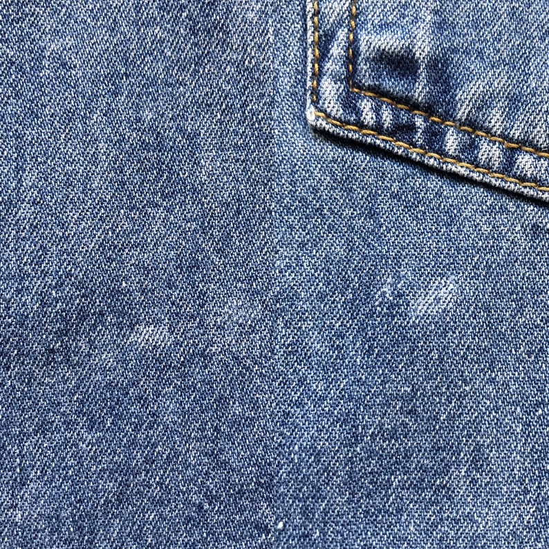 Pode incluir: Close-up de tecido denim azul com um canto de bolso. O denim tem uma apar&ecirc;ncia texturizada com detalhes de tecido vis&iacute;veis. O canto do bolso &eacute; refor&ccedil;ado com costuras amarelas, adicionando um elemento de contraste.