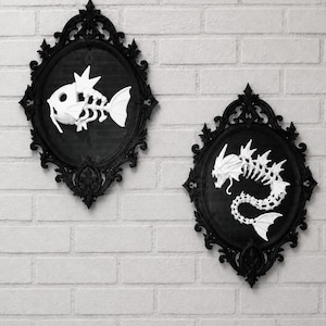 May include: Two oval black ornate frames containing white skeletal fish figures. The left frame displays a small fish skeleton with fins, while the right frame features a larger, elongated serpentine dragon-like skeleton with sharp head spikes.