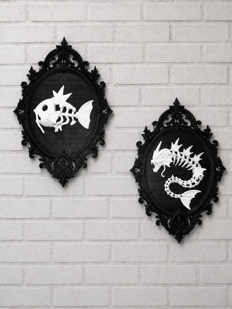 May include: Two oval black ornate frames containing white skeletal fish figures. The left frame displays a small fish skeleton with fins, while the right frame features a larger, elongated serpentine dragon-like skeleton with sharp head spikes.