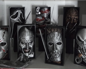 Puede incluir: Colección de máscaras de películas de terror exhibidas en cajas negras. Las máscaras incluyen Jason Voorhees, Ghostface, Pennywise, Michael Myers, Freddy Krueger, Jack Skellington y una Tortuga Ninja.