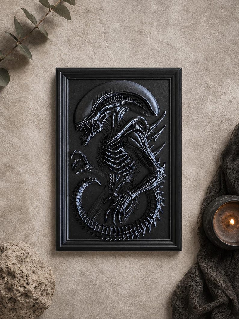 May include: A black framed relief sculpture of an Alien creature. The artwork features a detailed depiction of the creature's skeletal structure, elongated head, and tail. The sculpture is set against a neutral background, with a candle and fabric in the background.