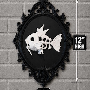 May include: A white fish skeleton relief mounted on a black oval background within an ornate black frame. The piece measures 12 inches high and 8 inches wide, featuring a stylized skeletal fish design with a crown-like fin on its head.