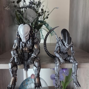 May include: Two detailed action figures sitting side by side. On the left, a Predator figure with a silver mask, dark armor, and dreadlocks. On the right, a black and silver Xenomorph figure with an elongated head and a long, curved tail.