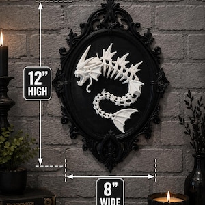 May include: A white skeletal sea creature sculpture mounted inside an ornate black oval frame. The wall art measures 12 inches high and 8 inches wide. The creature features a detailed skull, fins, and a curved spine, creating a stark contrast against the black background of the frame.