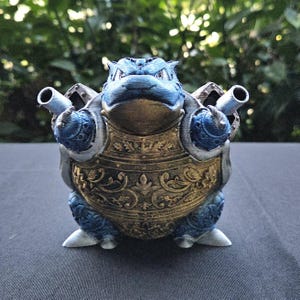 May include: A blue and gold figurine of a turtle-like creature with two cannons on its back. The figurine has intricate detailing on its shell and armor, and is set against a blurred green background.