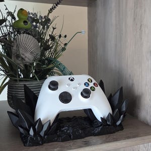 May include: A white Xbox controller rests on a black, crystal-like stand. The controller has black buttons and colored action buttons. The stand is on a wooden shelf, with a decorative plant arrangement in the background.