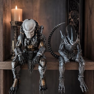 Puede incluir: Dos figuras detalladas sentadas una al lado de la otra. A la izquierda, un Predator con máscara plateada, armadura oscura y detalles de calaveras. A la derecha, un Xenomorfo negro con cabeza alargada lisa, torso estriado y cola larga.