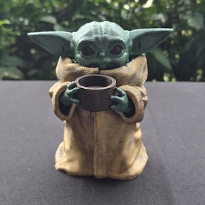 Könnte beinhalten: Grüne Figur einer Figur aus einem Science-Fiction-Franchise, die eine kleine braune Tasse hält. Die Figur trägt eine beige Robe. Die Figur hat große Ohren und ein detailliertes Gesicht. Der Hintergrund ist verschwommenes grünes Laub.