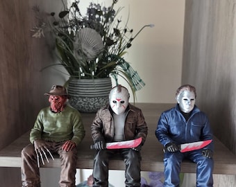 May include: Three horror movie character figurines sitting on a shelf. The left figure wears a green sweater and brown hat with metal claw fingers. The center figure wears a hockey mask and brown jacket holding a knife. The right figure wears a blue jumpsuit and white mask holding a knife.